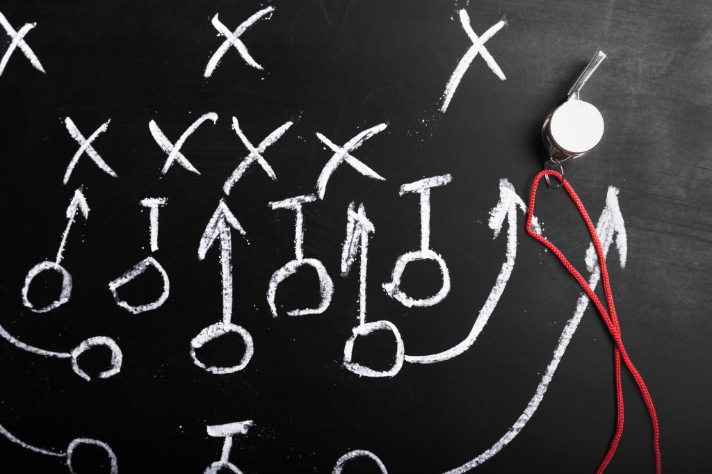 A football play diagram drawn in chalk on a blackboard with X’s, O’s, arrows, and a referee whistle on a red lanyard.