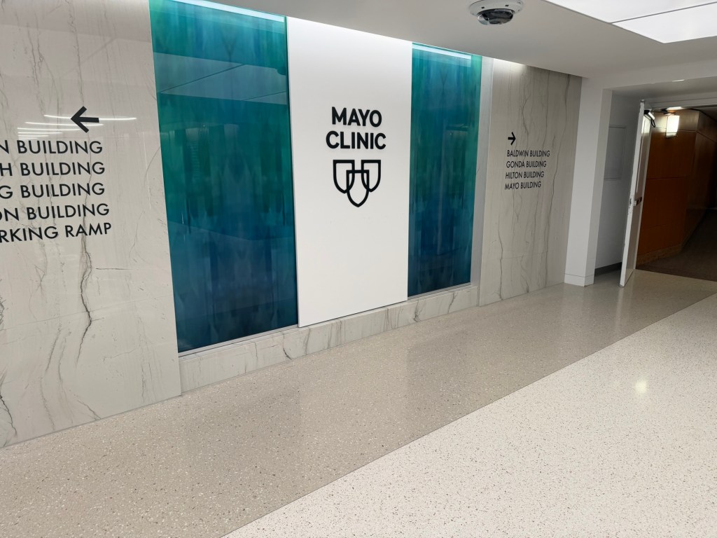 Hallway inside Mayo Clinic with a large white wall panel displaying the Mayo Clinic shield logo, flanked by blue-green glass panels and directional signs to the surrounding medical buildings.