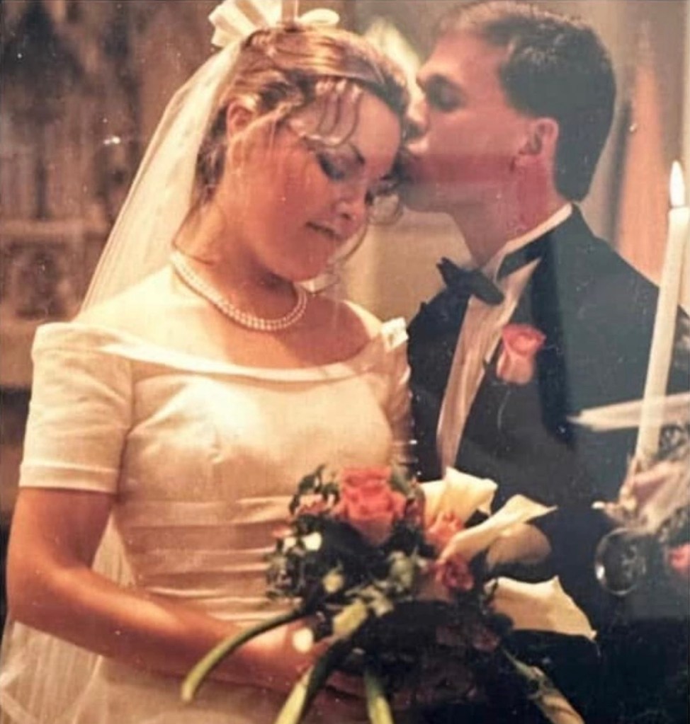 Elizabeth and Jeremy on their wedding day: Jeremy leans in and kisses Elizabeth on the forehead as she smiles softly, eyes closed. Elizabeth holds a bouquet of pink and white flowers, wearing a satin off-the-shoulder wedding gown, while Jeremy is in a classic black tuxedo with a white boutonniere. The moment is intimate, warm, and peaceful.