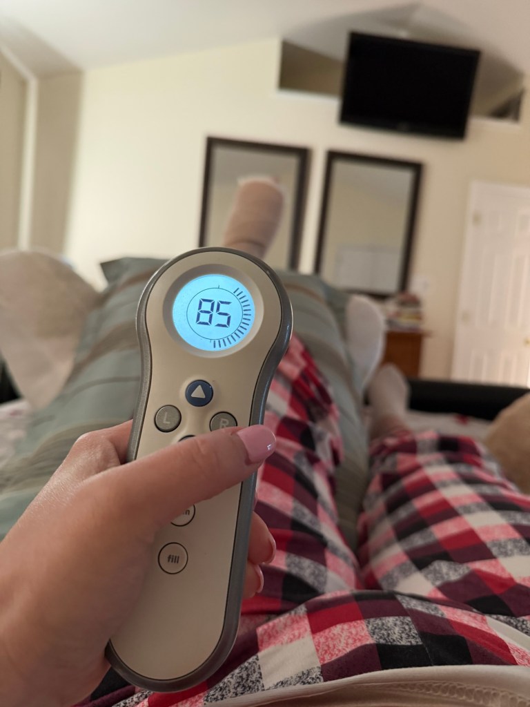 Hand holding a Sleep Number remote displaying 85 while resting on a bed with a bandaged foot elevated in the background.